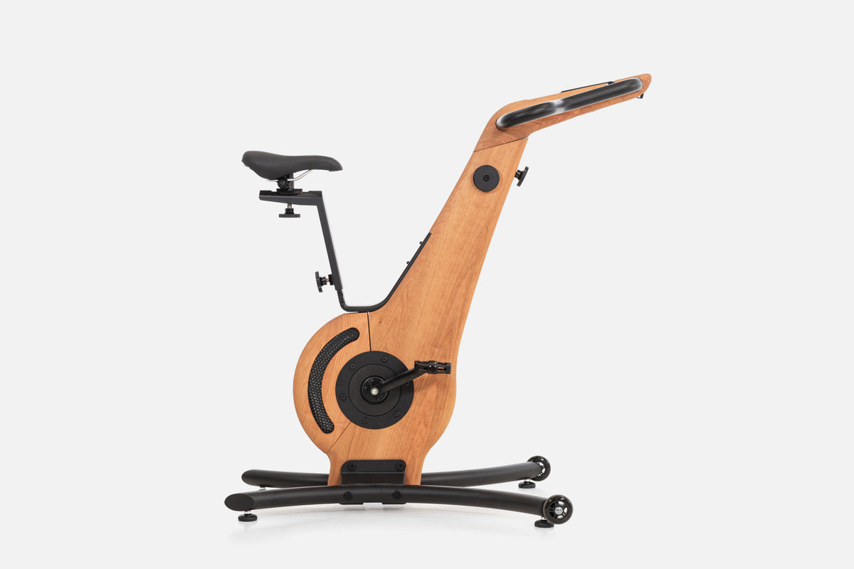 Side view of a warm golden-wood indoor cycle with a black saddle, mesh flywheel cover, and a handlebar touchscreen