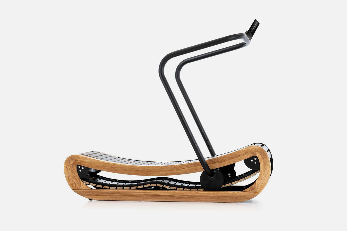 Side view of a curved manual treadmill with a pale wood frame, black slats, and a raised black handlebar