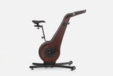 Side view of a dark reddish-brown wooden indoor cycle with a black saddle, mesh flywheel cover, and a handlebar touchscreen