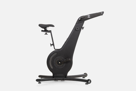 Side view of a black wooden indoor cycle with a black saddle, mesh flywheel cover, and a handlebar touchscreen