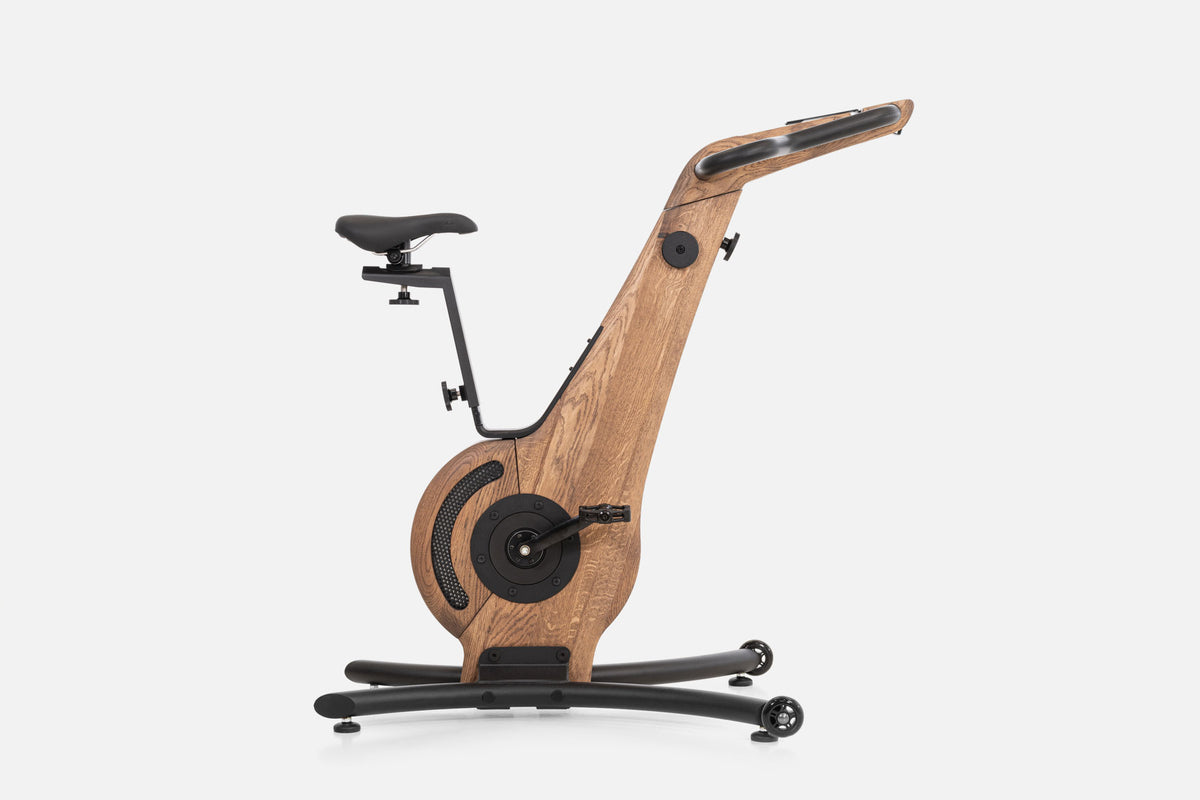 Side view of a medium-brown wooden indoor cycle with a black saddle, mesh flywheel cover, and a handlebar touchscreen