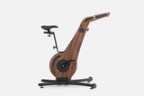 Side view of a dark brown wooden indoor cycle with a black saddle, mesh flywheel cover, and a handlebar touchscreen