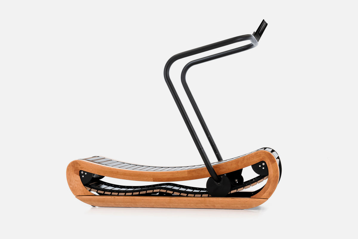 Side view of a curved manual treadmill with a warm golden wood frame, black slats, and a raised black handlebar